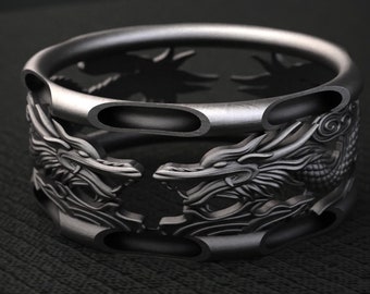 Könnte beinhalten: Ein dunkelgrauer Metallring mit einem durchbrochenen Design, bei dem sich zwei Drachenköpfe gegenüberstehen. Das Band hat eine zylindrische, ausgehöhlte Struktur mit glatten, abgerundeten Kanten und detaillierten, erhabenen Drachenschuppen.