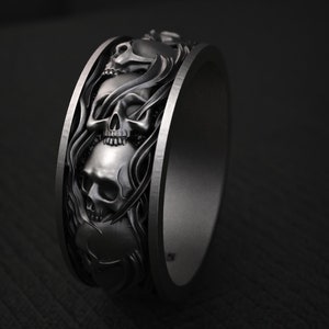 May include: A dark gray metal ring featuring a continuous relief pattern of multiple human skulls intertwined with flowing, smoke-like or ribbon-like decorative elements that wrap around the entire outer circumference of the band.