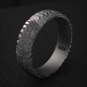 May include: A dark grey sterling silver ring with a textured, granular surface and a row of raised, rectangular segments along one edge. The interior of the band is smooth and clearly stamped with the hallmark 925, indicating sterling silver content.