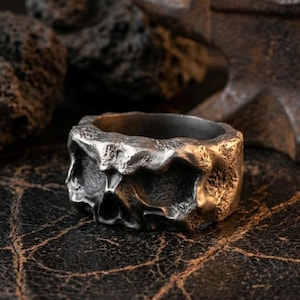 May include: A silver-toned metal ring with a rugged, uneven surface texture and a dark, oxidized finish. The band features deep, irregular indentations and a heavy, cast appearance, showcasing a raw and weathered aesthetic throughout the entire piece.