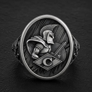 Op de afbeelding: Een zilverkleurige ring met een ovaalvormig blad met een reliëf van een Spartaanse krijger met een helm en een zwaard. Onder de krijger bevindt zich een ruitvormig embleem met de letter C. De achtergrond heeft een textuur met lijnen.