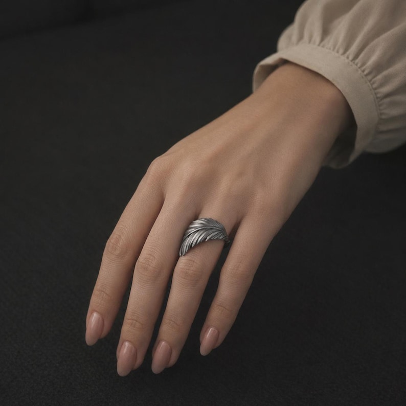 K&ouml;nnte beinhalten: Ein silberfarbener Ring in Form einer Feder, der an einem Finger getragen wird. Der Ring hat detaillierte Federtexturen und ein geschwungenes Design. Der dunkle Hintergrund hebt den metallischen Glanz des Rings hervor. Der Ring ist ein Schmuckst&uuml;ck.