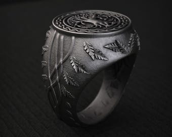 May include: A dark gray metal ring featuring a circular top with an embossed tree design surrounded by a braided border. The band is decorated with raised leaf patterns and intertwined vine-like lines, showcasing a textured, matte finish throughout.