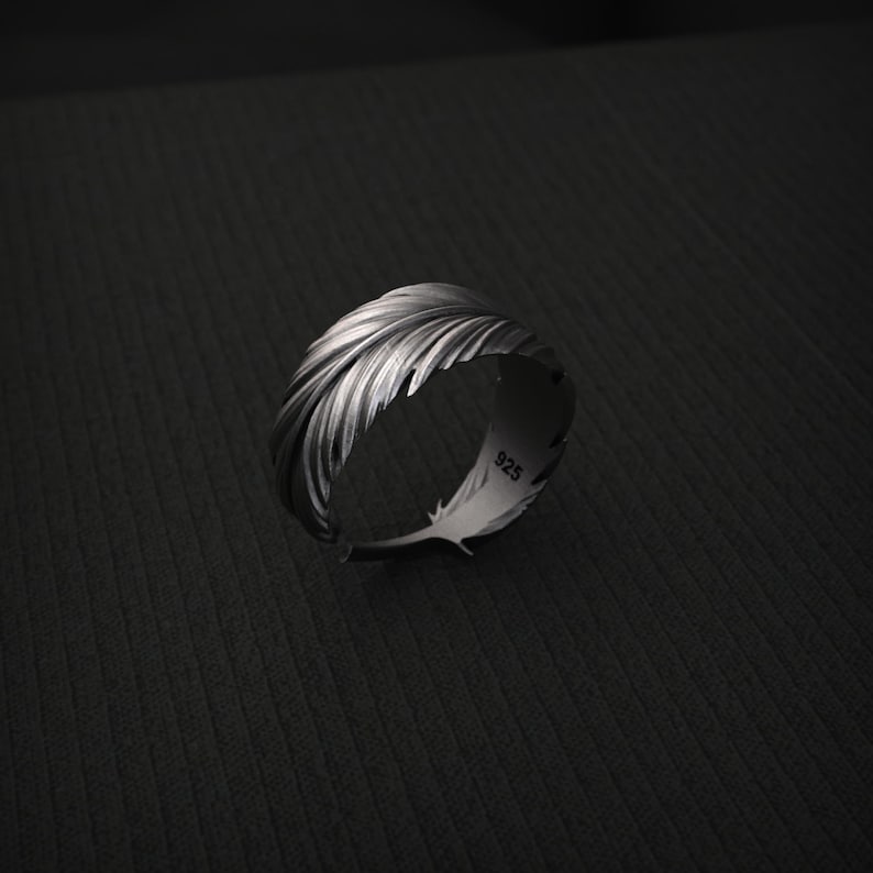 K&ouml;nnte beinhalten: Ein silberner Ring mit einem Federdesign. Der Ring ist mit einzelnen Federborsten detailliert und hat einen 925-Stempel auf der Innenseite. Der Ring ist vor einem dunklen Hintergrund platziert.