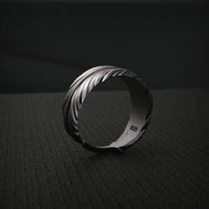 K&ouml;nnte beinhalten: Ein silberner Ring mit einem strukturierten Federdesign. Der Ring besteht aus dunklem, oxidiertem Metall und tr&auml;gt die Nummer "925" auf der Innenseite. Der Ring ist vor einem dunklen Hintergrund platziert.
