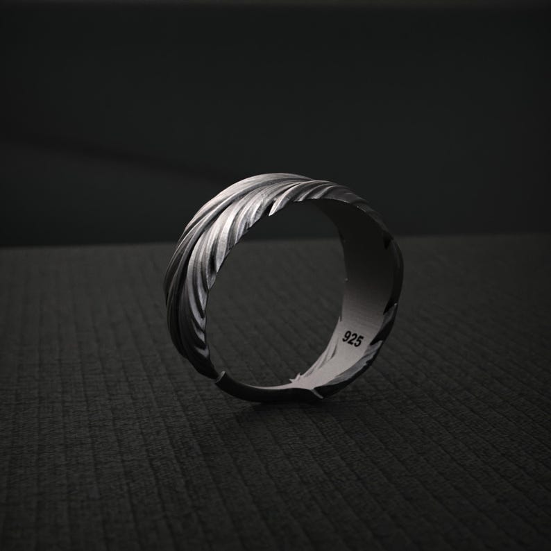 K&ouml;nnte beinhalten: Ein silberner Ring mit einem strukturierten Federdesign. Der Ring besteht aus dunklem, oxidiertem Metall und tr&auml;gt die Nummer "925" auf der Innenseite. Der Ring ist vor einem dunklen Hintergrund platziert.