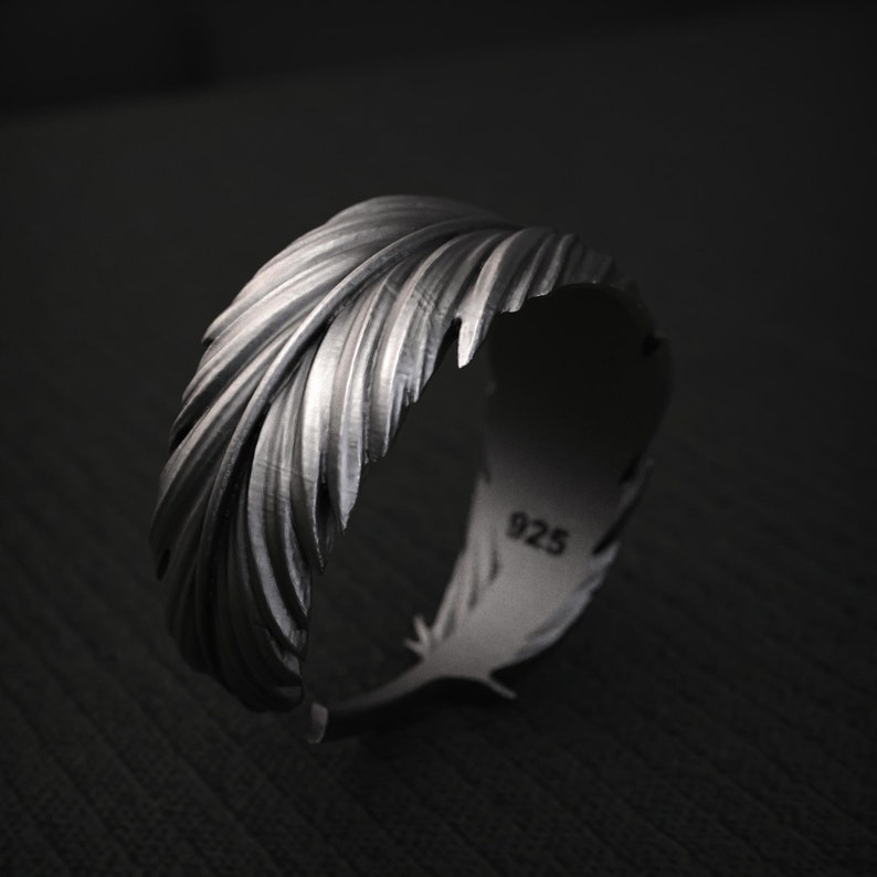 K&ouml;nnte beinhalten: Ein silberner Ring in Form einer Feder. Der Ring hat detaillierte, strukturierte Linien und ist innen mit "925" markiert. Der Ring ist vor einem dunklen Hintergrund platziert.