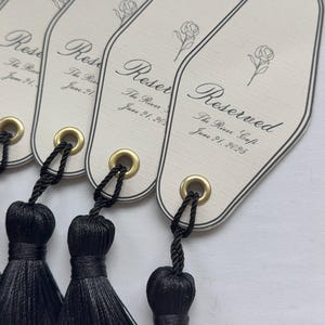 Peut inclure: &Eacute;tiquettes allong&eacute;es de couleur cr&egrave;me avec du texte noir et une illustration de rose. Chaque &eacute;tiquette est orn&eacute;e d'un pompon noir fix&eacute; par un &oelig;illet dor&eacute;. Le texte indique "Reserved" et "The River Cafe, June 21, 2025".