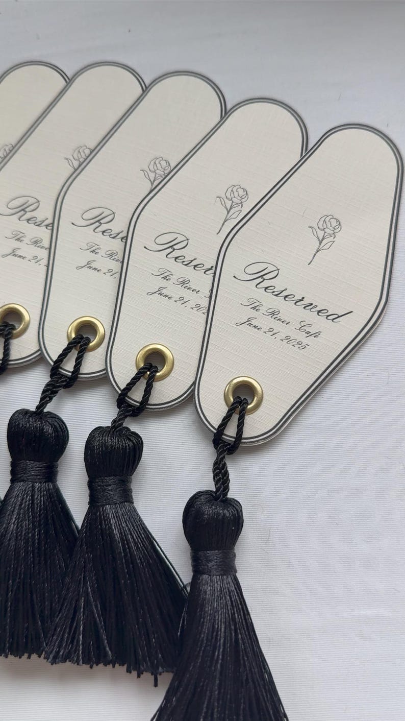 Peut inclure: &Eacute;tiquettes allong&eacute;es de couleur cr&egrave;me avec du texte noir et une illustration de rose. Chaque &eacute;tiquette est orn&eacute;e d'un pompon noir fix&eacute; par un &oelig;illet dor&eacute;. Le texte indique "Reserved" et "The River Cafe, June 21, 2025".