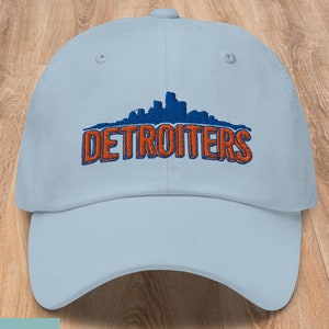 May include: A light blue baseball cap with an embroidered skyline and the text "DETROITERS" in orange and blue.