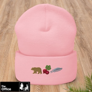 Puede incluir: Gorro de punto rosa con un oso blanco, una remolacha roja y un avión blanco bordados en la parte delantera. El gorro tiene un dobladillo.