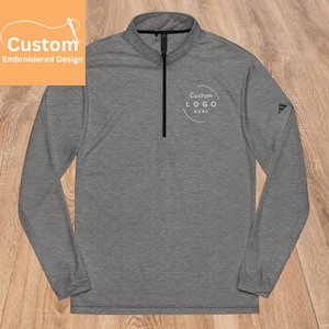 Puede incluir: Un jersey de rendimiento gris de manga larga con una cremallera negra y un logotipo circular blanco que dice "Custom LOGO HERE" en el pecho izquierdo.