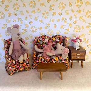 May include: Two plush toy mice sitting on floral patterned chairs with a wooden coffee table in between. The chairs and table are miniature sized and the background is a floral wallpaper.