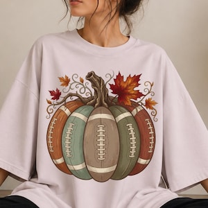 May include: A light pink short-sleeve t-shirt featuring a large graphic of a pumpkin constructed from segments that resemble brown and green footballs. The design includes a textured brown stem and several orange and red autumn maple leaves.