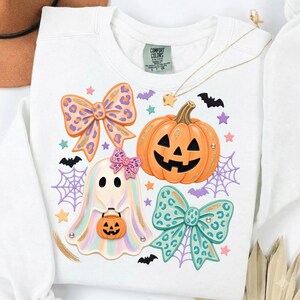 May include: A white sweatshirt featuring a graphic print of a jack-o'-lantern, a ghost holding a pumpkin bucket, two leopard print bows, spiderwebs, bats, and stars. The inner neck label reads Comfort Colors with size S.