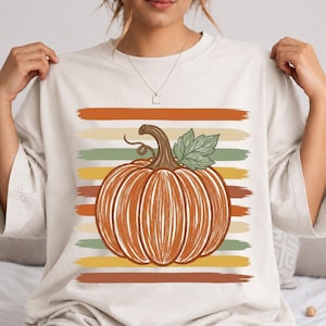 May include: A cream-coloured t-shirt featuring a central graphic of an orange pumpkin with a brown stem and a green leaf. The pumpkin is layered over a series of horizontal, distressed-style stripes in shades of orange, beige, and muted green.