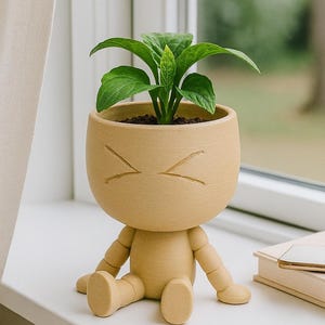 Może przedstawiać: Kapryśna, jasnobrązowa doniczka w kształcie siedzącej postaci z okrągłą głową i kątowymi oznaczeniami oczu. Doniczka zawiera żywą zieloną roślinę z dużymi liśćmi. Postać siedzi na białym parapecie.
