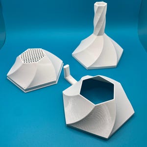 Puede incluir: Tres objetos blancos, geométricos, impresos en 3D se muestran sobre un fondo azul. Uno tiene una parte superior perforada, otro tiene un tallo retorcido y el tercero tiene una forma hexagonal abierta con un pequeño asa. Los objetos tienen un diseño moderno y abstracto.
