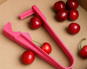 May include: A pink cherry pitter tool and several ripe red cherries are arranged inside a cardboard box. The pitter is made of plastic and has a long handle with a forked end for removing cherry pits. The cherries are glossy and fresh.