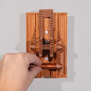 Puede incluir: Una cubierta de interruptor de luz de color cobre, impresa en 3D, con un diseño de engranajes mecánicos. La cubierta es rectangular con una serie de engranajes y pistas. Una mano gira el interruptor cilíndrico. El diseño recuerda la estética steampunk.