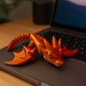 Könnte beinhalten: Eine detaillierte, orange und rote Drachenfigur mit beweglichen Gelenken und Flügeln, die auf einem Laptop liegt. Der Drache hat einen strukturierten Körper und ist mit erhobenem Kopf dargestellt. Die Figur ist etwa 15 cm lang.