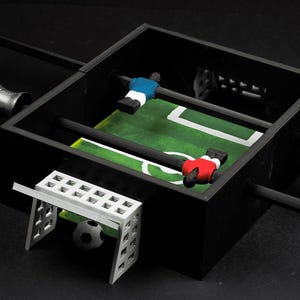 Puede incluir: Una mesa de futbolín en miniatura con marco negro, campo de juego verde y marcas blancas. Dos equipos de jugadores con camisetas azules y rojas están colocados en las barras. Una portería blanca y un balón de fútbol en primer plano.
