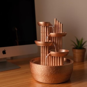 以下が含まれることがあります： 複数のレベルから水が流れ落ちる銅色の屋内噴水。 噴水は、テクスチャ加工されたベースと、一連の垂直の柱とボウルを備えています。 小さな鉢植えの植物とコンピューターが背景にあります。