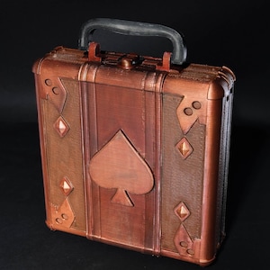Pode incluir: Uma maleta cor de cobre com uma alça preta. A frente apresenta um grande símbolo de espadas e detalhes em forma de diamante. A maleta tem uma aparência texturizada e uma forma retangular, sugerindo um design vintage ou temático.