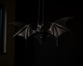 Op de afbeelding: Een donkere metalen vleermuissculptuur met uitgespreide vleugels hangt aan een dunne draad. De vleermuis heeft gedetailleerde vleugelpatronen en een getextureerd lichaam. De achtergrond is donker.