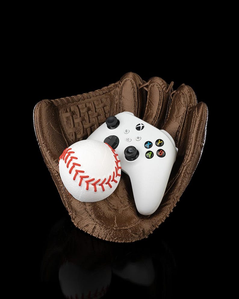 May include: A baseball glove holds a white baseball with red stitching and a white Xbox controller. The glove is brown with detailed stitching. The controller has black buttons and colored action buttons.