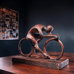 May include: A bronze sculpture of a cyclist in a racing position. The figure is atop a bicycle with circular wheels, mounted on a rectangular base. The sculpture has a textured finish and is displayed on a wooden surface.