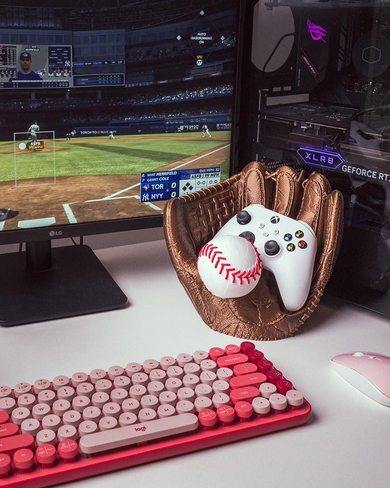 May include: A baseball-themed gaming setup featuring a baseball glove-shaped holder with a baseball and a white controller. A red and pink keyboard and a pink mouse are in the foreground. A computer monitor displays a baseball game.