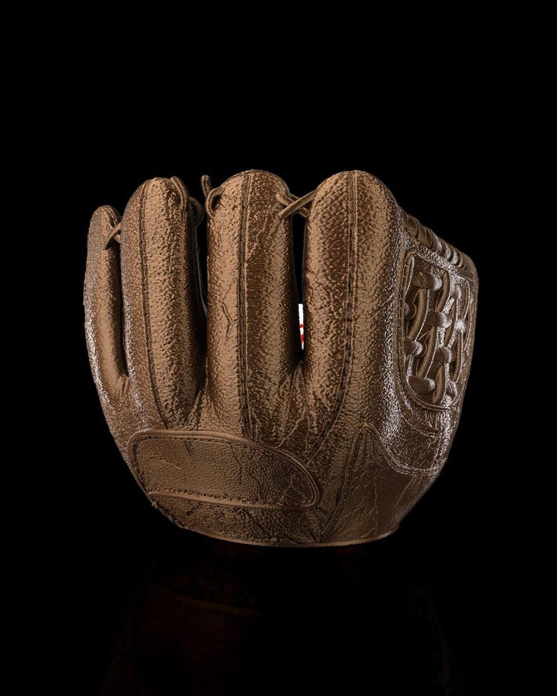 May include: A brown baseball glove with a textured surface. The glove has a classic design with visible stitching and a decorative emblem on the side. The image is set against a black background, highlighting the glove's details.