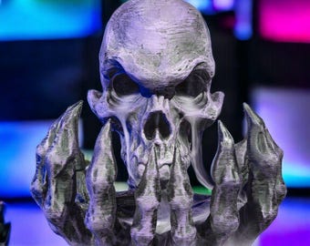 May include: A detailed, gray skull sculpture with outstretched hands, emanating white vapor. The skull has a textured surface and is set against a blurred, colorful background. The sculpture appears to be 3D-printed.