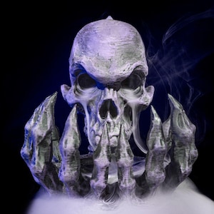 Puede incluir: Una calavera gris detallada con dientes afilados y una superficie texturizada, sostenida por manos esqueléticas. Volutas de humo blanco rodean la calavera sobre un fondo oscuro, creando una imagen espeluznante con temática de Halloween.