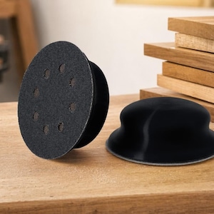 Puede incluir: Dos discos de lijado negros y una base de lijado negra se muestran sobre una superficie de madera. El disco de lijado redondo tiene múltiples agujeros y una superficie texturizada. La base de lijado tiene un diseño curvo.