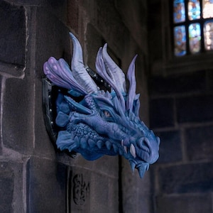 May include: A blue dragon head wall sculpture with purple highlights. The dragon features sharp teeth and textured scales, mounted on a black shield. The sculpture is set against a dark, textured wall, creating a fantasy theme.