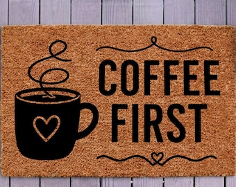 Op de afbeelding: Een rechthoekige bruine kokosmat met een zwarte afbeelding van een koffiemok met een hartje en stoom, vergezeld van de zwarte tekst COFFEE FIRST. De mat ligt op een lichtpaarse houten veranda vloer voor een donkerpaarse deur.