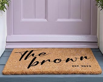 Op de afbeelding: Een rechthoekige, beige kokosmat met zwarte tekst in scriptstijl die The brown leest, samen met de tekst EST. 2023. De mat ligt voor een lichtpaarse deur met een koperen brievenbus en twee witte plantenbakken met witte en paarse bloemen.