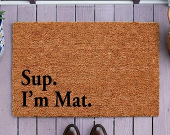 Op de afbeelding: Een rechthoekige bruine kokosmat met de zwarte tekst Sup. I'm Mat. aan de linkerkant. De mat heeft een grove, vezelige textuur en ligt op een lichtpaarse houten veranda voor een donkerpaarse deur.