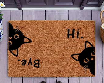 Op de afbeelding: Een rechthoekige bruine kokosmat met zwarte illustraties van twee kattenkoppen die uit tegenovergestelde hoeken kijken. De tekst "Hi." staat in het zwart rechtsboven en de tekst "Bye." staat in het zwart linksonder op de mat.