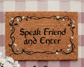 Op de afbeelding: Een rechthoekige bruine kokosmat met een zwarte rankenrand met kleine witte bloemen en de tekst Speak Friend and Enter in een gestileerd zwart lettertype, gecentreerd op een roze en wit geblokte stoffen mat.