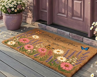 Op de afbeelding: Een rechthoekige kokosmat met een kleurrijke bloemenprint van roze en witte bloemen, groen blad en vlinders. De tekst "Home Sweet Home" en "Est. 2024" is in zwarte letters in het midden van de mat gedrukt.