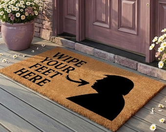 Op de afbeelding: Een rechthoekige beige kokosmat met het zwarte silhouet van een hoofd en schouders. De tekst "WIPE YOUR FEET HERE" staat in zwarte hoofdletters gedrukt boven een pijl die naar rechts naar het silhouet wijst.