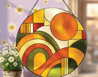 Op de afbeelding: Een ronde glas-in-lood zonnevanger met een abstract geometrisch landschapsontwerp, voorzien van een grote oranje zon, gebogen heuvels in tinten geel, oranje en groen, en een zwart metalen frame opgehangen aan een dunne metalen ketting.