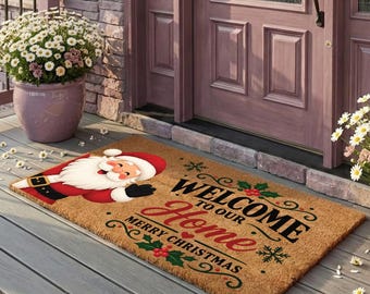 Może przedstawiać: Prostokątna wycieraczka z włókna kokosowego z ilustracją Świętego Mikołaja po lewej stronie oraz napisem "WELCOME TO OUR Home MERRY CHRISTMAS" czcionką w kolorze czarnym i czerwonym. Mata ozdobiona jest zielonymi liśćmi ostrokrzewu i czerwonymi jagodami.