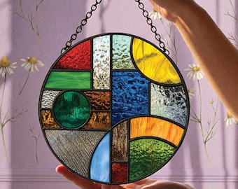 Op de afbeelding: Een ronde glas-in-lood zonnevanger met een geometrische indeling van veelkleurige glaspanelen in rood, blauw, groen, geel en oranje, bijeengehouden door donkere metalen profielen en opgehangen aan een zwarte metalen ketting.