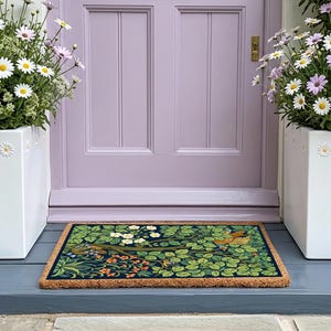 May include: A rectangular doormat with a nature-inspired design featuring birds, leaves, and flowers in shades of green, orange, and white. The mat is placed in front of a lilac door, flanked by white flower pots with daisies.