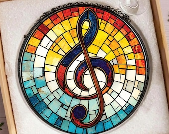 Op de afbeelding: Een ronde glas-in-lood zonnevanger met een centraal vioolsleutelsymbool. Het ontwerp heeft een mozaïekpatroon met levendige segmenten van rood, oranje, geel, lichtblauw en donkerblauw glas, gevat in een dunne zwarte metalen omlijsting.