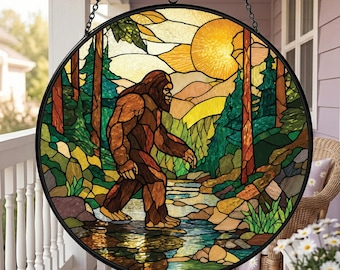 Op de afbeelding: Een ronde glas-in-lood zonnevanger met een bruine Bigfoot-figuur die door een bos met een beekje loopt, tegen een achtergrond van een grote gele zon. Het object is omlijst met zwart metaal en hangt aan een zwarte ketting.